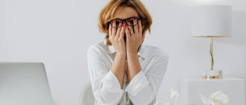 Stresszes nő ül az irodában, arcát a kezébe temeti, befejezetlen munkafeladattal küzd.